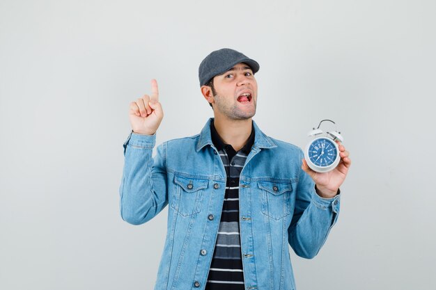 Hombre joven apuntando hacia arriba mientras sostiene el reloj en la chaqueta, gorra y parece hablador. vista frontal.