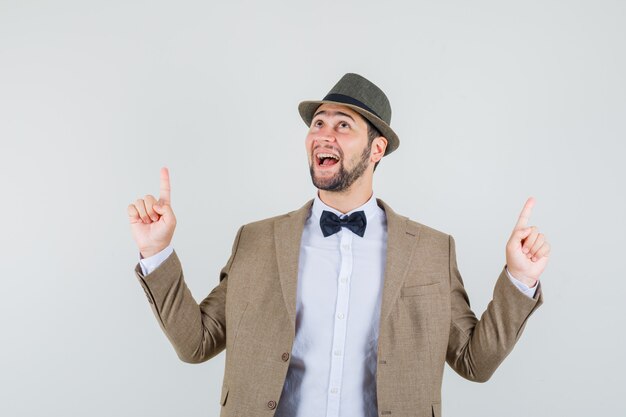 Hombre joven apuntando hacia arriba en traje, sombrero y mirando alegre, vista frontal.