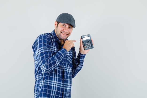 Hombre joven apuntando a la calculadora en camisa, gorra y mirando alegre. vista frontal.