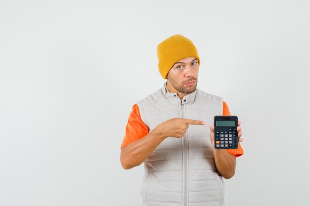 Hombre joven apuntando a la calculadora en camiseta, chaqueta, sombrero y mirando serio. vista frontal.