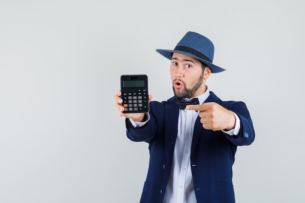 Hombre joven apuntando a la calculadora en traje, sombrero y mirando confiado, vista frontal.