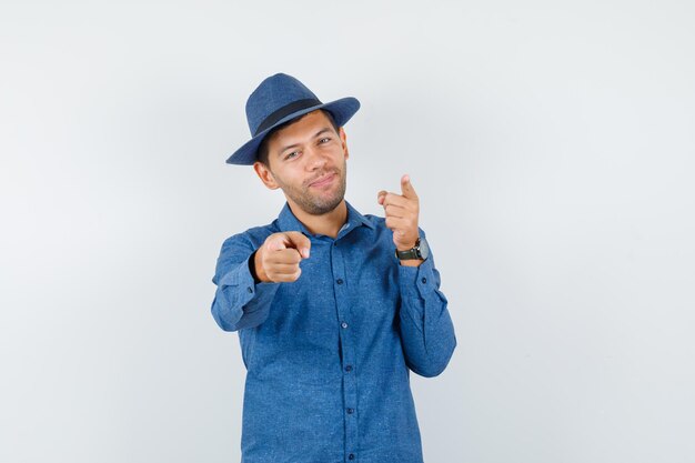 Hombre joven apuntando a la cámara con camisa azul, sombrero y mirando alegre. vista frontal.