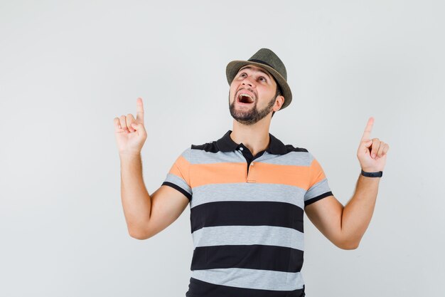 Hombre joven apuntando con el dedo hacia arriba en camiseta, sombrero y mirando optimista, vista frontal.