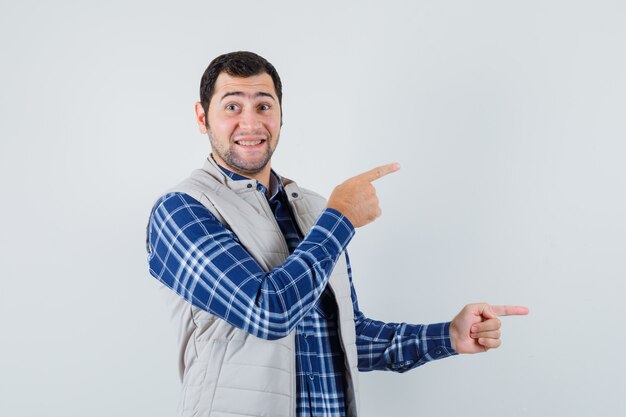 Hombre joven apuntando a un lado en camisa, chaqueta sin mangas y mirando contento. vista frontal.