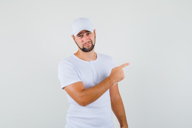 Hombre joven apuntando a un lado con camiseta blanca, gorra y mirando insatisfecho. vista frontal.