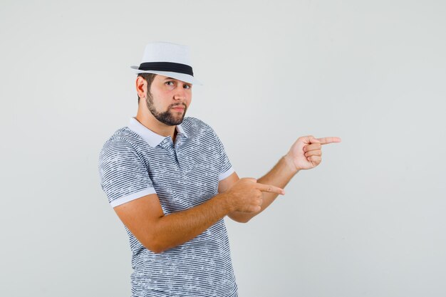 Hombre joven apuntando a un lado con camiseta a rayas, sombrero y aspecto lindo. vista frontal.