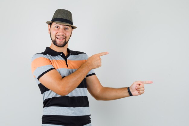 Hombre joven apuntando hacia el lado en camiseta, sombrero y mirando alegre.