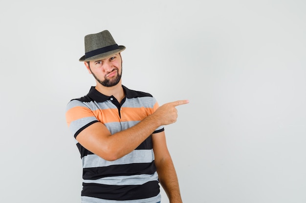 Hombre joven apuntando hacia el lado en camiseta, sombrero y mirando dudoso.