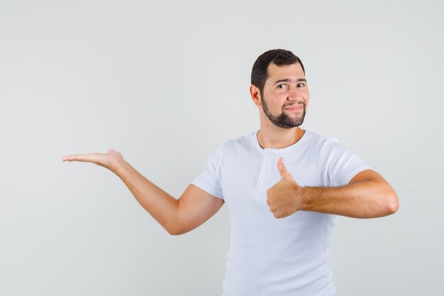 Hombre joven apuntando a un lado mientras muestra el pulgar hacia arriba con una camiseta blanca y parece contento. vista frontal.