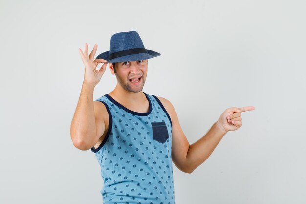 Hombre joven apuntando hacia un lado, sosteniendo el sombrero en camiseta azul, sombrero y mirando alegre. vista frontal.