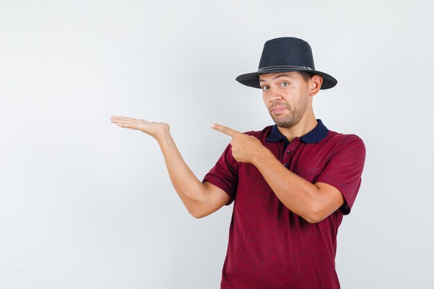 Hombre joven apuntando hacia otro lado mientras extiende la palma en camiseta, sombrero y mirando vacilante. vista frontal.