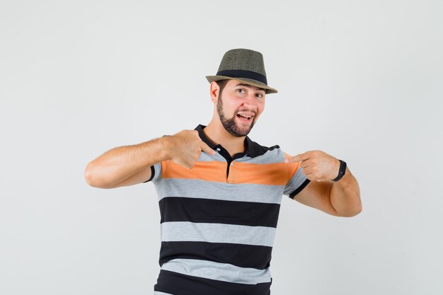 Hombre joven apuntando a sí mismo en camiseta, sombrero y mirando orgulloso. vista frontal.