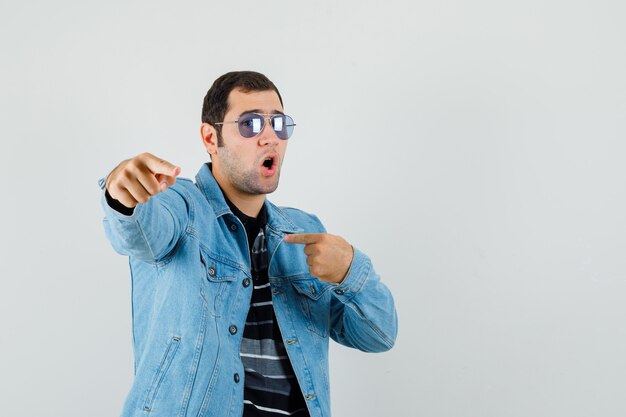 Hombre joven apuntando a sí mismo y a ti en camiseta, chaqueta y mirando confiado