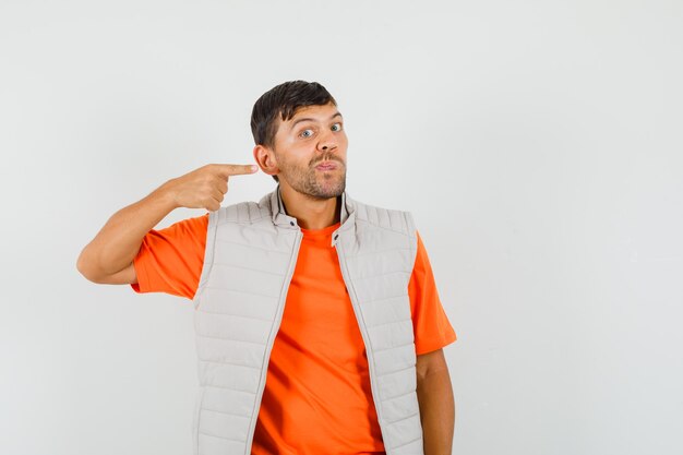 Hombre joven apuntando a su oreja en camiseta, chaqueta y mirando extraño, vista frontal.