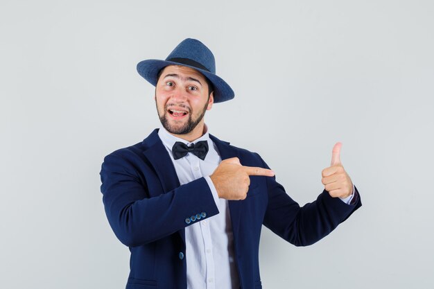 Hombre joven apuntando a su pulgar hacia arriba en traje, sombrero y mirando alegre, vista frontal.