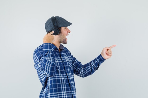 Hombre joven apuntando a sus auriculares y a un lado en camisa, gorra y mirando alegre, vista frontal.