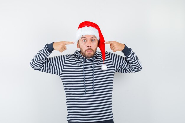 Hombre joven apuntando a sus ojos con capucha, gorro de Papá Noel y mirando desconcertado.