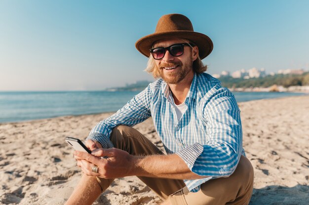 Hombre joven atractivo hipster sentado en la playa por el mar en vacaciones de verano