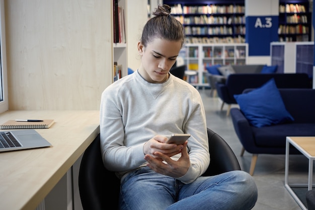 Hombre joven atractivo en jeans y suéter leyendo sms en su teléfono inteligente