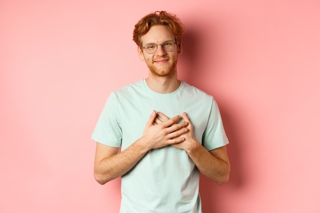 Hombre joven atractivo con el pelo pelirrojo cogidos de la mano en el corazón y sonriendo agradecido diciendo gracias e ...