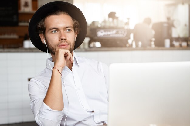 Hombre joven atractivo que sostiene la mano en la barbilla, que tiene una mirada pensativa, que se sienta frente a una computadora portátil abierta con auriculares y escucha un audiolibro en línea