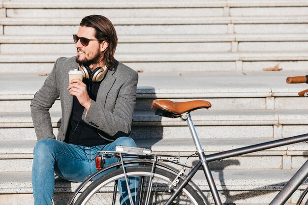 Un hombre joven atractivo que sostiene la taza de café para llevar que se sienta en las escaleras cerca de la bicicleta