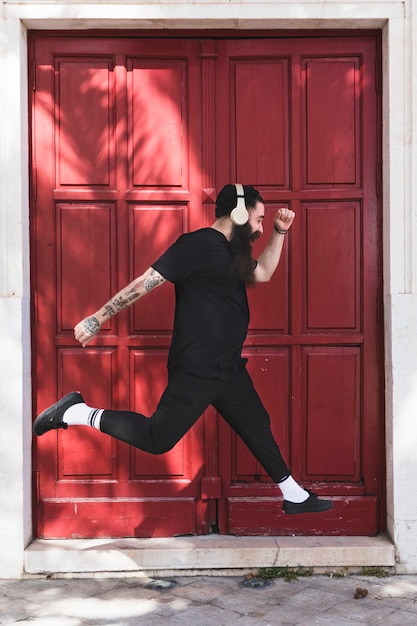 Foto gratuita hombre joven con el auricular en su cabeza que salta contra puerta cerrada roja