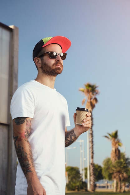 Hombre joven con barba y tatuajes en camiseta blanca sin etiqueta con una taza de café contra el cielo azul y palmeras