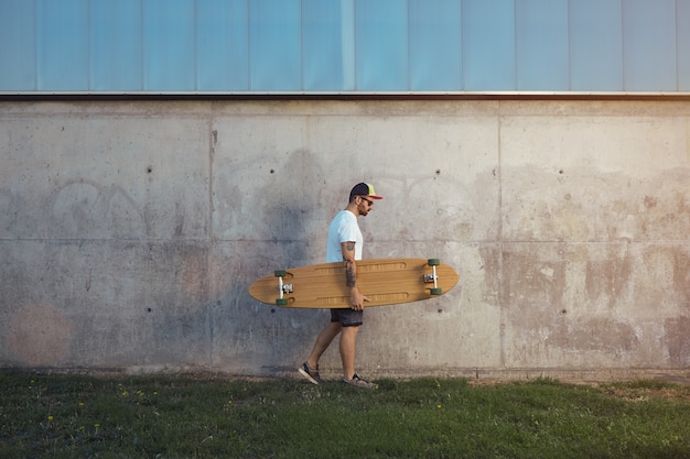 Foto gratuita hombre joven con barba y tatuajes vistiendo una camiseta blanca lisa, pantalones cortos, zapatillas y gorra de béisbol llevando un longboard a lo largo de una pared de hormigón