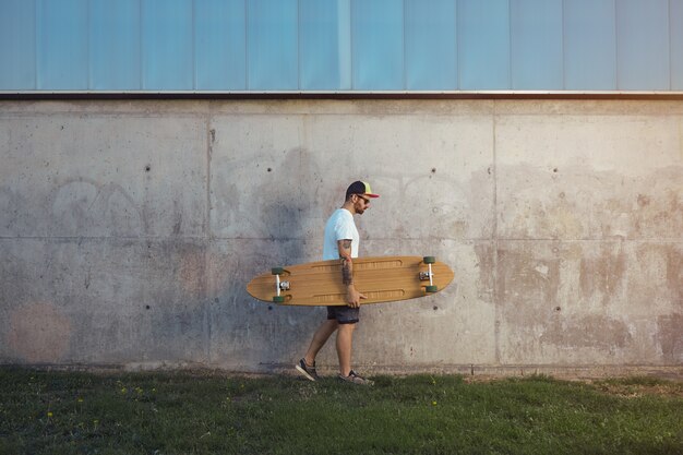 Hombre joven con barba y tatuajes vistiendo una camiseta blanca lisa, pantalones cortos, zapatillas y gorra de béisbol llevando un longboard a lo largo de una pared de hormigón