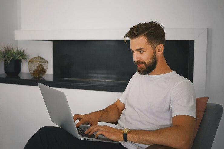 Foto gratuito hombre joven barbudo elegante que se relaja en casa con una computadora portátil en su regazo, mecanografía mientras envía mensajes en línea con una chica interesante a través del sitio web de citas, con una expresión facial curiosa y alegre