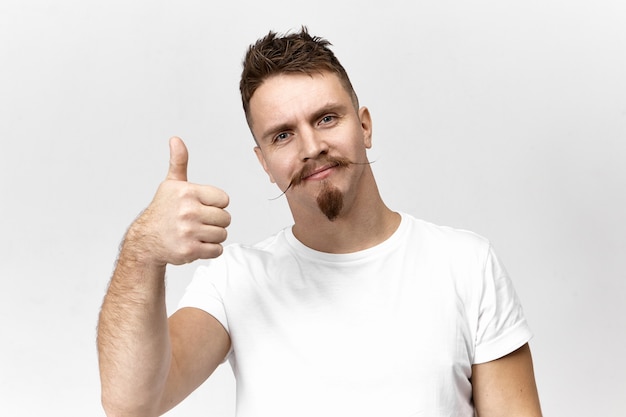 Hombre joven barbudo con estilo con bigote de manillar posando en estudio en camiseta casual blanca sonriendo con alegría, haciendo gesto de pulgar hacia arriba, aprobando una buena película. Concepto de positividad y aprobación