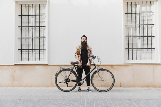 Hombre joven barbudo de pie con bicicleta contra la pared