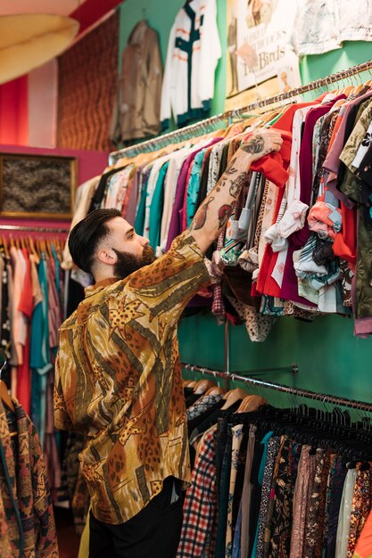 Hombre joven barbudo que elige la camiseta roja del carril en la tienda de ropa