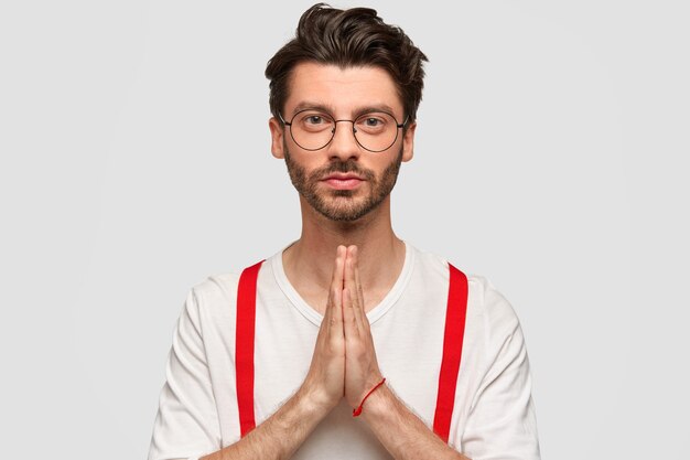 Foto gratuita hombre joven barbudo seguro de sí mismo, confiado, mantiene las manos en gesto de oración, viste un suéter blanco con tirantes rojos, tiene rostro serio, cree en lo mejor. joven atractivo tiene fe en lo mejor