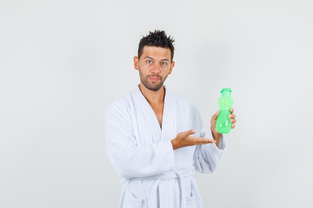Hombre joven en bata de baño blanca mostrando una botella de agua de plástico, vista frontal.