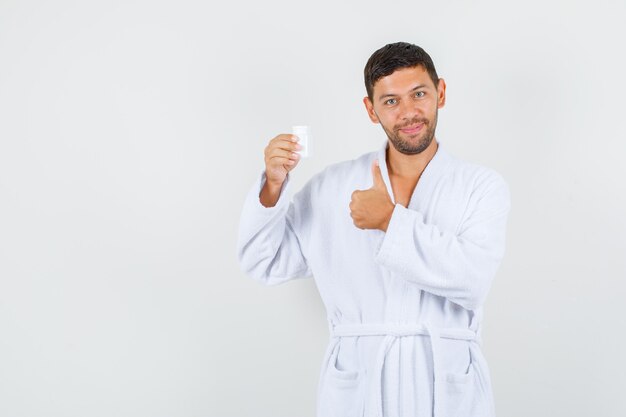 Hombre joven en bata de baño blanca sosteniendo el frasco de medicina con el pulgar hacia arriba y mirando alegre, vista frontal.
