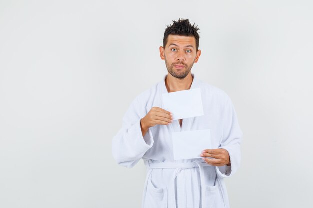 Hombre joven en bata de baño blanca sosteniendo hojas de papel en blanco, vista frontal.