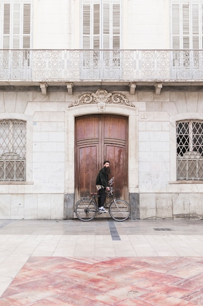 Hombre joven con la bicicleta que se coloca delante del edificio del vintage