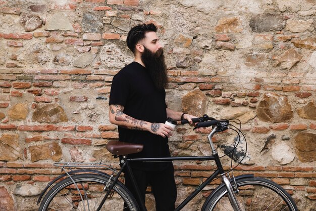 Hombre joven con la bicicleta que sostiene la taza de café disponible en la mano que se coloca cerca de la pared resistida