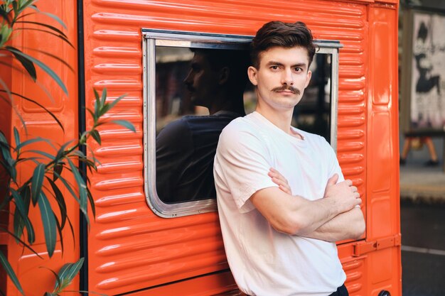 Hombre joven con bigote en una camiseta blanca apoyado en una furgoneta roja cogidos de la mano mientras mira soñadoramente a la cámara en la calle de la ciudad