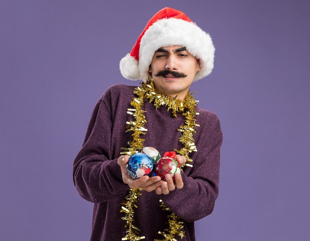 Hombre joven bigotudo con gorro de navidad de santa con oropel alrededor de su cuello sosteniendo bolas de navidad con una sonrisa escéptica de pie sobre la pared púrpura