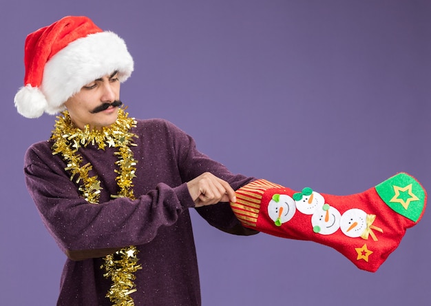 Foto gratuita hombre joven bigotudo con gorro de papá noel de navidad con oropel alrededor de su cuello sosteniendo una media de navidad mirando confundido de pie sobre fondo púrpura