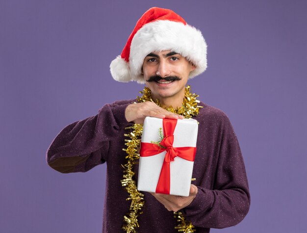 Hombre joven bigotudo con gorro de Papá Noel de Navidad con oropel alrededor de su cuello sosteniendo el regalo de Navidad con cara feliz sonriendo de pie sobre la pared púrpura