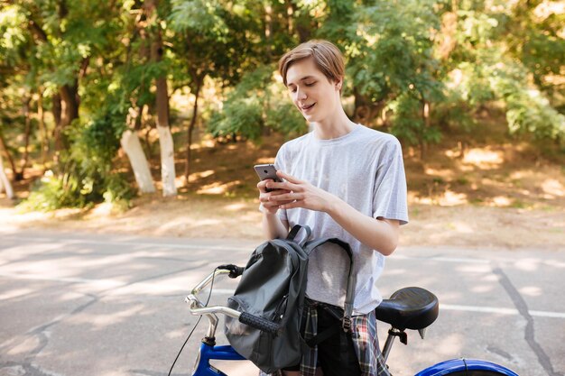 Hombre joven con cabello rubio de pie con mochila y bicicleta en el parque Retrato de niño sonriente fresco de pie y usando su teléfono celular mientras pasa tiempo en el parque