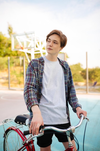 Hombre joven con cabello rubio de pie en pantalones cortos y camisa casual con bicicleta roja en la cancha de baloncesto Retrato de niño pensativo sosteniendo bicicleta y mirando soñadoramente a un lado