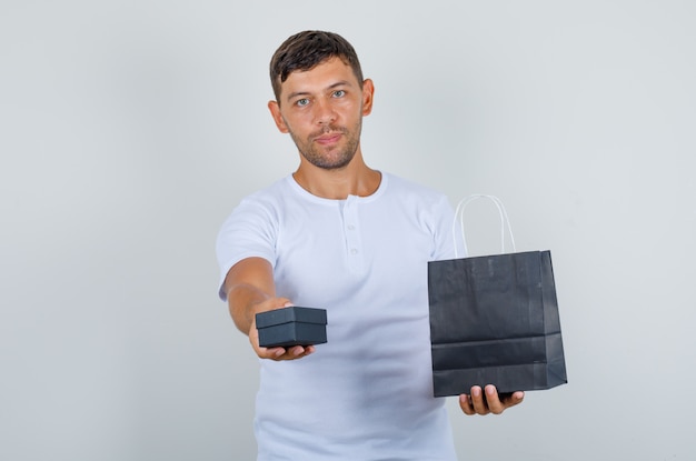 Hombre joven con caja actual y bolsa de papel en camiseta blanca, vista frontal.