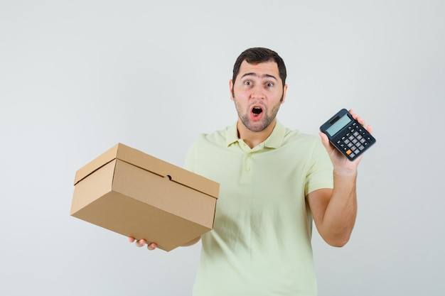 Hombre joven con caja de cartón y calculadora en camiseta y mirando asombrado. vista frontal.