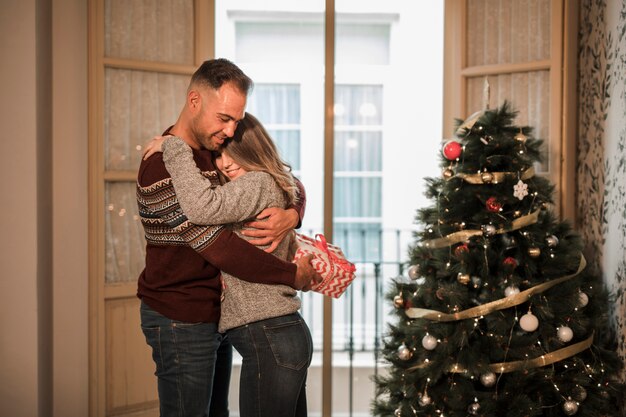 Hombre joven con la caja de regalo que abraza a la mujer alegre cerca del árbol de navidad