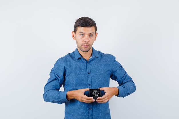 Hombre joven con caja de reloj en camisa azul y mirando sensible, vista frontal.